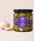 A jar of Divina Garlic Stuffed Olives Greece on a light beige wooden surface next to a whole garlic bulb and two garlic cloves. The jar has a black lid and a purple circular label.