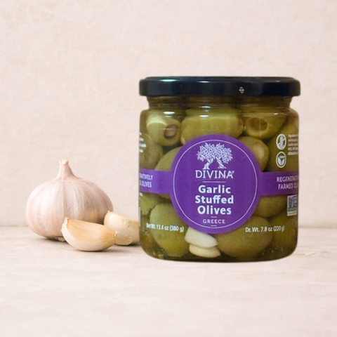 A jar of Divina Garlic Stuffed Olives Greece on a light beige wooden surface next to a whole garlic bulb and two garlic cloves. The jar has a black lid and a purple circular label.